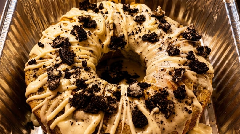 Bundt cake with white frosting and crushed Oreo cookies in a foil pan.