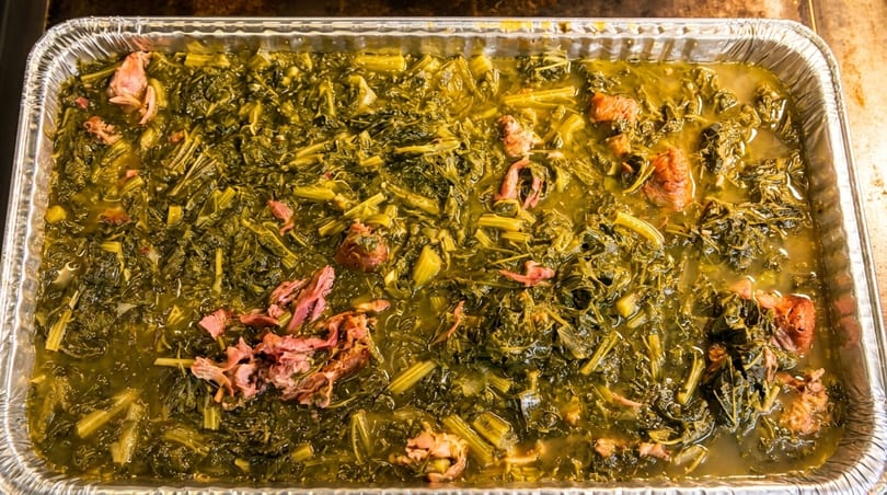 Cooked collard greens with ham hocks in a large aluminum pan.